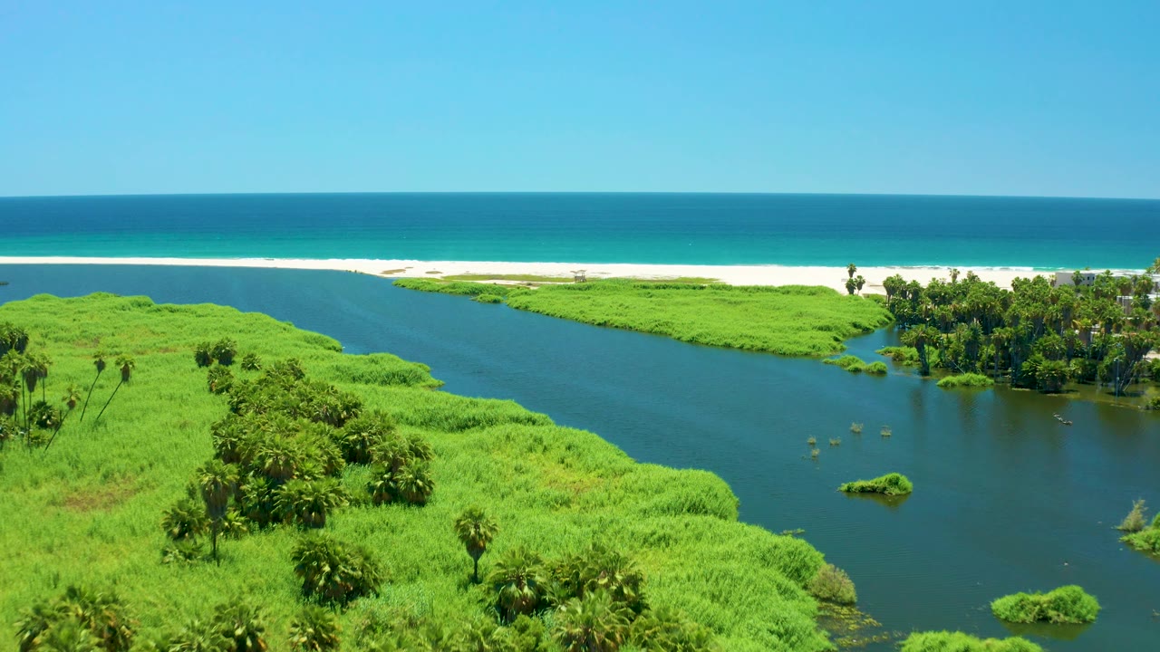 Aerial view of the San Jose Del Cabo Estuary with the river bend framed by lush vegetation in the foreground, beach and ocean in the background, and no visible people.