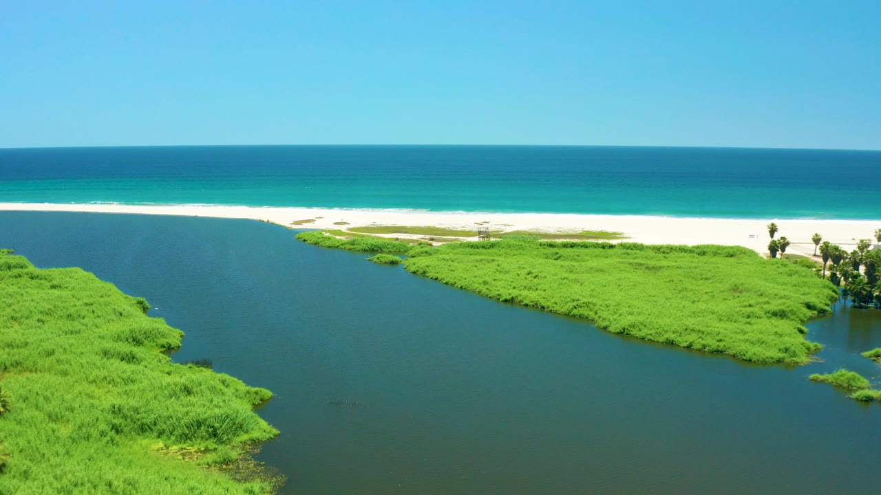 Opening aerial frame over San Jose del Cabo Estuary with dark river water and bright green vegetation in the foreground, white sand and surf beyond, blue ocean on the horizon, and no visible people.