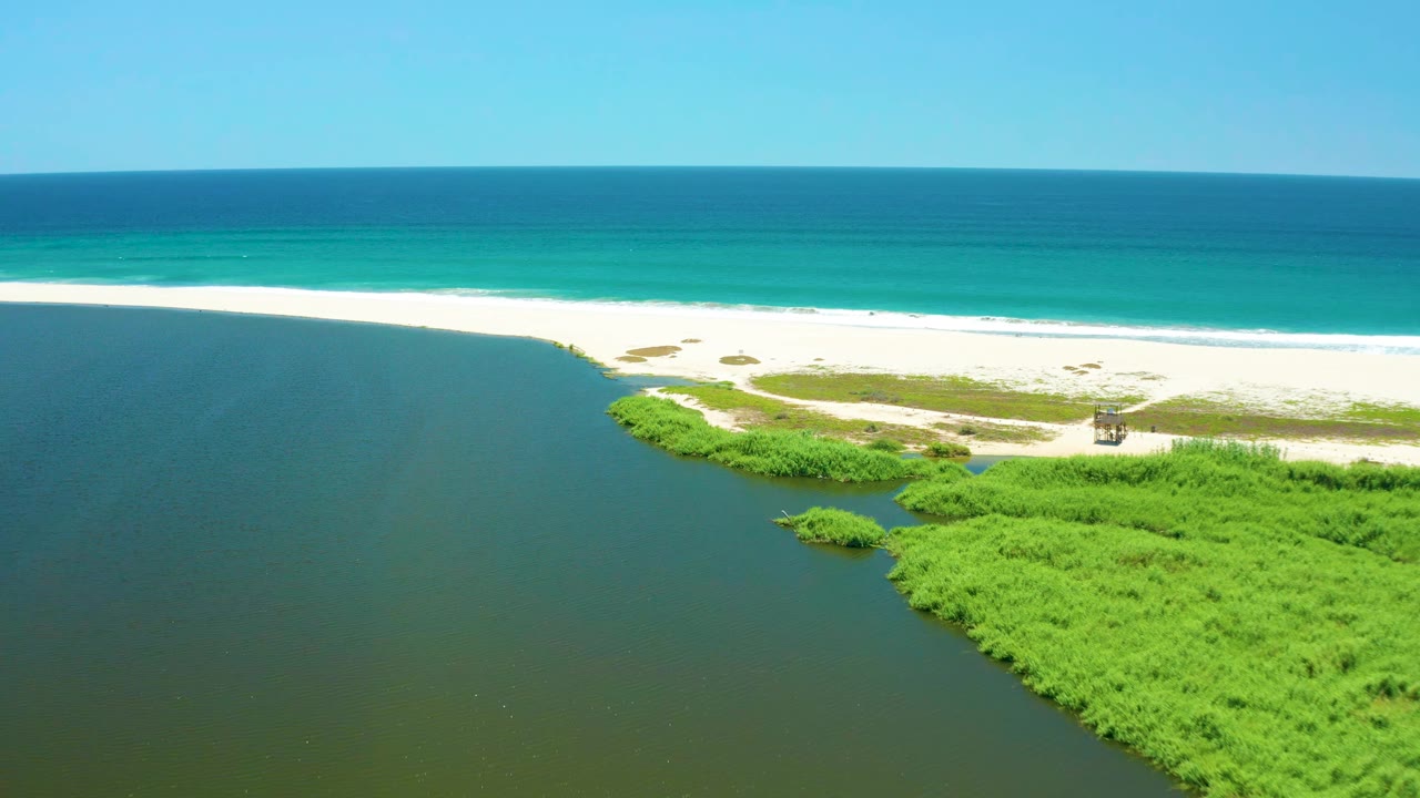 Aerial view of San Jose Del Cabo Estuary with lagoon water filling the left side, green vegetation along the lower shore, white sand and turquoise ocean across the top, and no visible people.