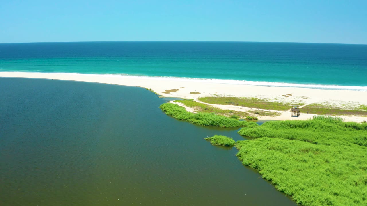 Aerial frame over San Jose Del Cabo Estuary showing more of the inlet curve, green vegetation patches, pale sand, a small beach structure near the right side, turquoise ocean, and no visible people.