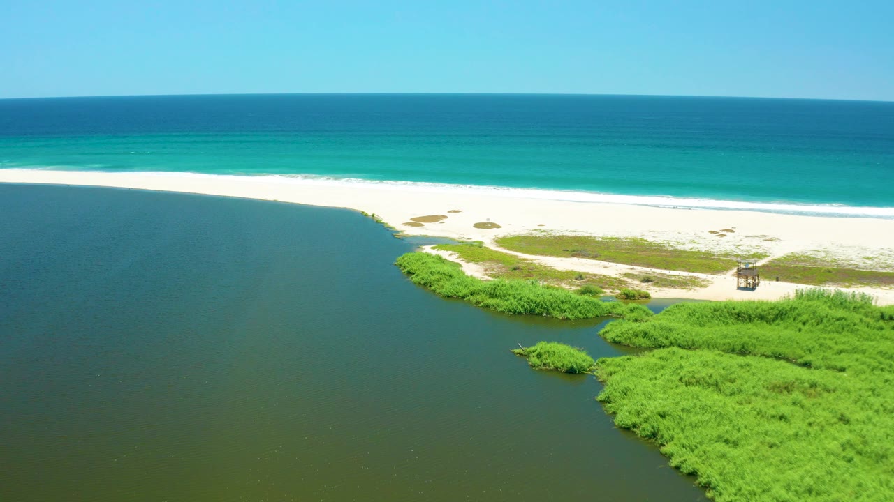 San Jose Del Cabo Estuary aerial still with dark lagoon water occupying the lower left, a pale sandbar and green vegetation in the center, turquoise ocean at the top, and no visible people.