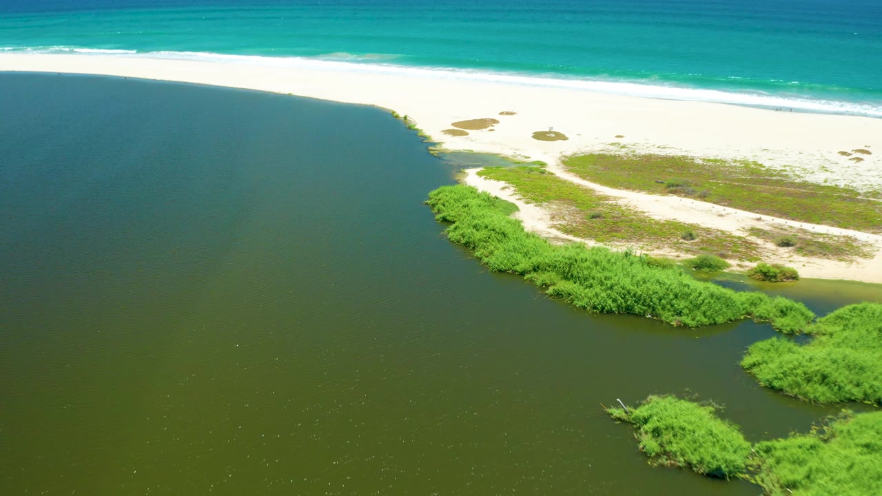 Drone-height frame of San Jose del Cabo Estuary with a wide area of dark river water, a white sand barrier, small dune scrub patches, ocean surf at the top edge, and no visible people.