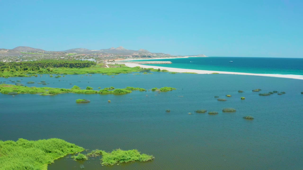 At one second, the San Jose del Cabo Estuary remains framed from above with marsh patches across the lagoon, a long white sand beach separating lagoon from ocean, distant hills, clear sky, and no visible people.