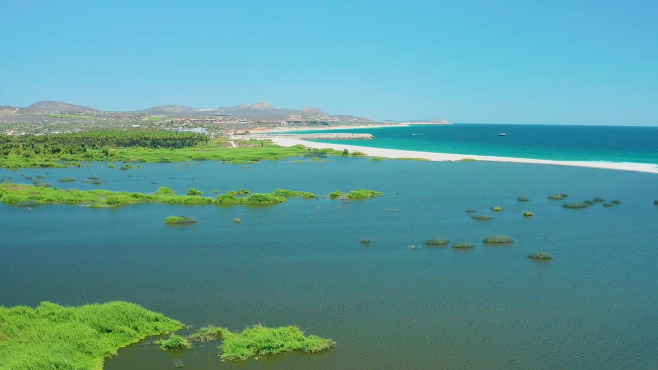 At two seconds, the aerial view shows scattered green vegetation floating in shallow San Jose del Cabo Estuary water, with the sandbar and ocean stretching across the upper frame, hills beyond, and no visible people.