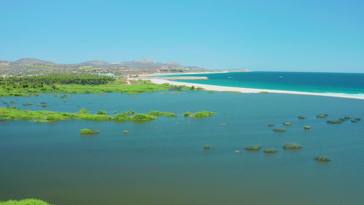Opening aerial frame over San Jose Del Cabo Estuary, with green lagoon water in front, scattered vegetation patches, a pale beach strip, blue ocean, distant hills, and no visible people.
