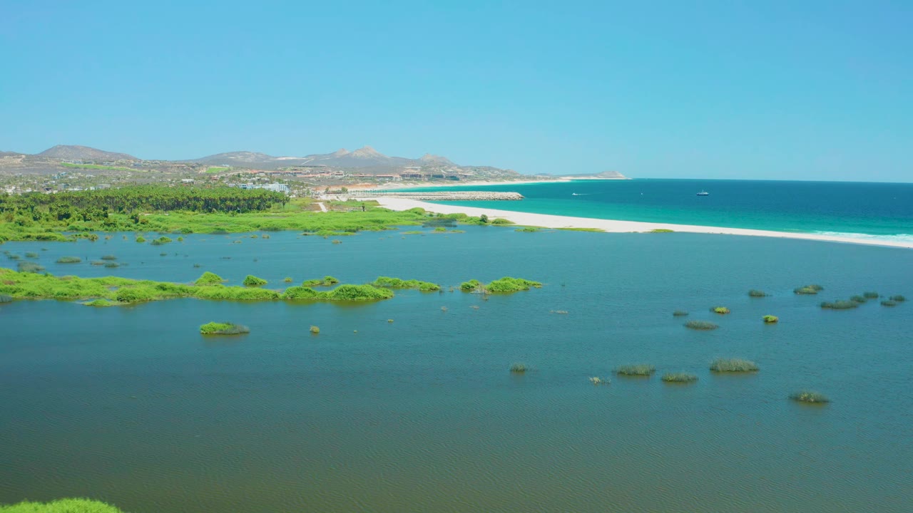 Coastal lagoon with vegetation patches, beach, ocean, hills, and clear sky.
