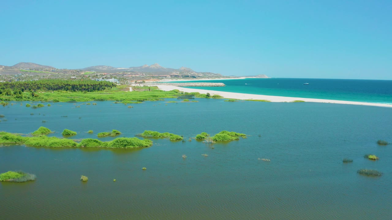 A wide aerial moment over San Jose Del Cabo Estuary places small green vegetation islands across shallow water, with the sandy beach and darker blue ocean stretching behind, and no visible people.