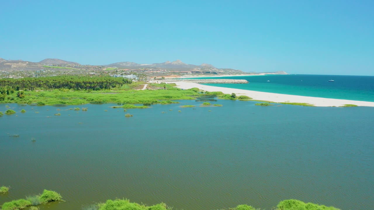 Aerial view of lagoon water, wetland greenery, sandy beach, ocean, hills, and clear sky.