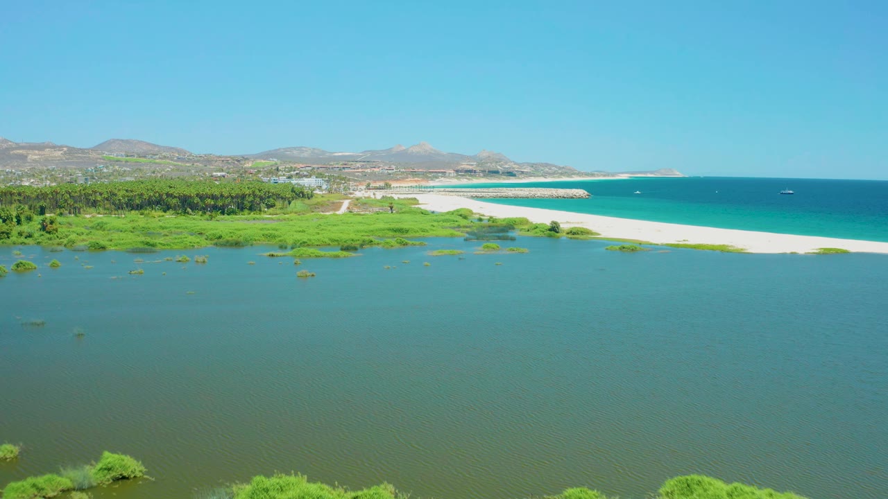 Wide aerial view at the San Jose del Cabo Estuary showing calm green-blue lagoon water, wetland plants, a bright sandbar, open ocean to the right, distant hills, clear sky, and no visible people.
