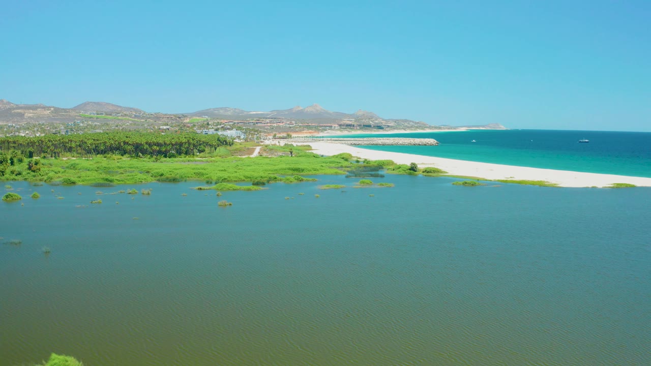 Aerial frame of the San Jose del Cabo Estuary with textured lagoon water below, patches of wetland vegetation near shore, a long sandy beach, blue ocean, low hills, clear sky, and no visible people.