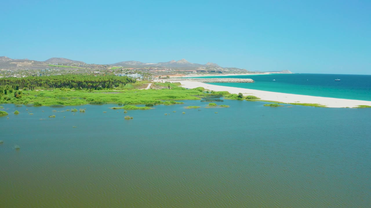 Mid-clip aerial view over the San Jose del Cabo Estuary, with calm water filling the lower frame, dense green vegetation left of center, pale beach and ocean beyond, distant hills, clear sky, and no visible people.