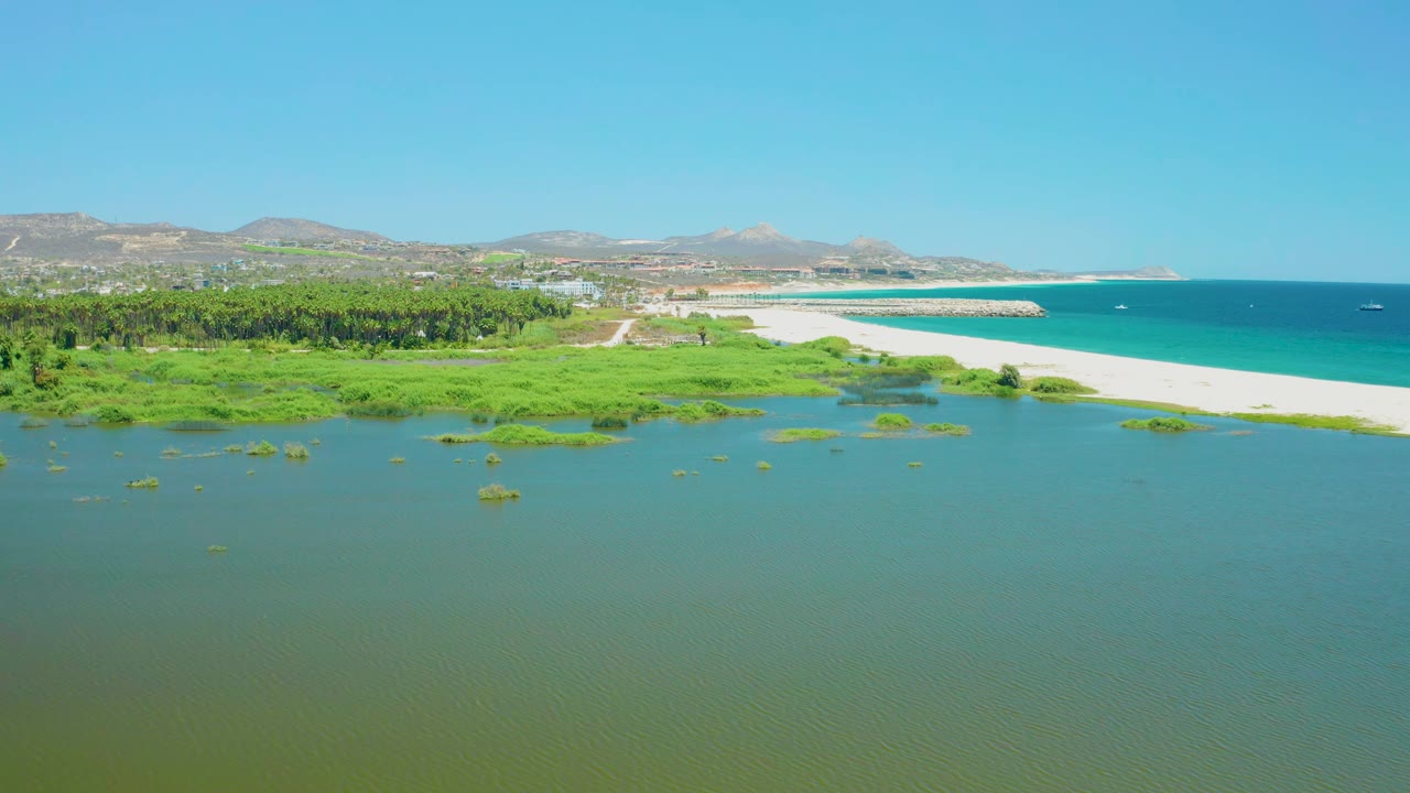 Wide aerial of coastal wetlands, lagoon water, sandy beach, turquoise ocean, and distant mountains.