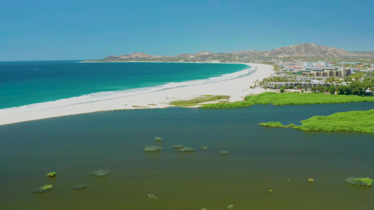 San Jose del Cabo Estuary appears from a high aerial angle with scattered algae patches across the foreground lagoon, a curved white beach, blue ocean, right-side coastal buildings, distant hills, and no visible people.