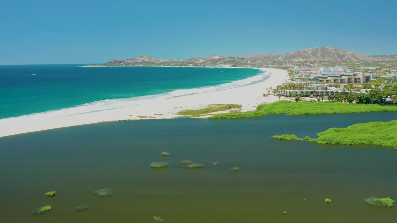 Mid-clip aerial view over San Jose del Cabo Estuary showing floating green algae in the lagoon foreground, a long white beach, turquoise ocean water, buildings grouped near the right shoreline, low hills, and no visible people.
