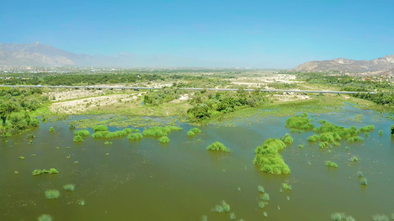 Aerial frame from San Jose del Cabo Estuary showing green bushes scattered through floodwater, a horizontal road in the midground, distant buildings, mountains, and no visible people.