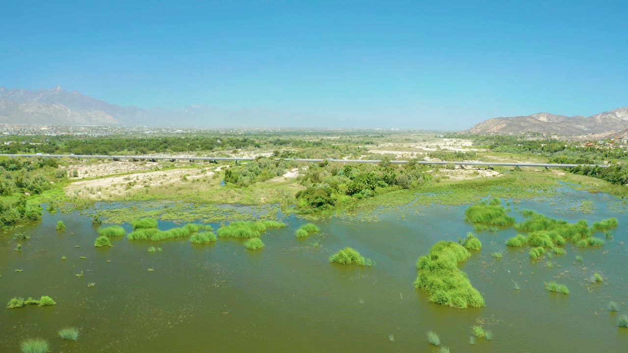 San Jose del Cabo Estuary aerial frame with green scrub and reeds emerging from floodwater, the road stretching behind the marsh, distant buildings, hazy mountain ridges, and no visible people.