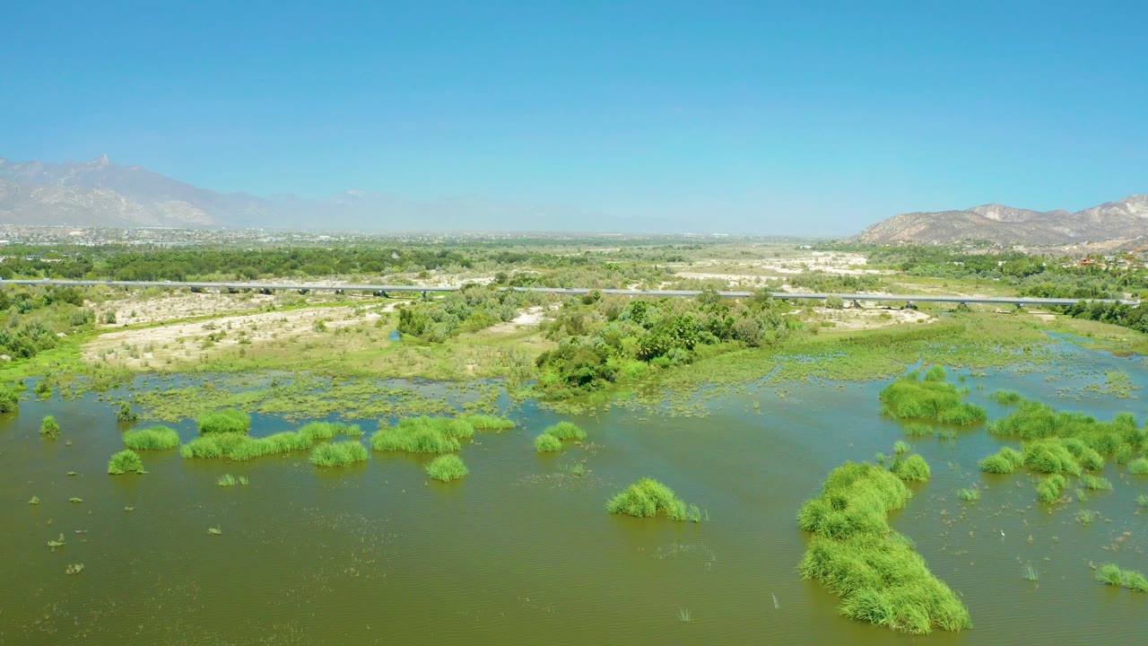 Opening aerial view of San Jose del Cabo Estuary with green wetland water in the foreground, a thin highway line in the midground, distant mountains, and no visible people.