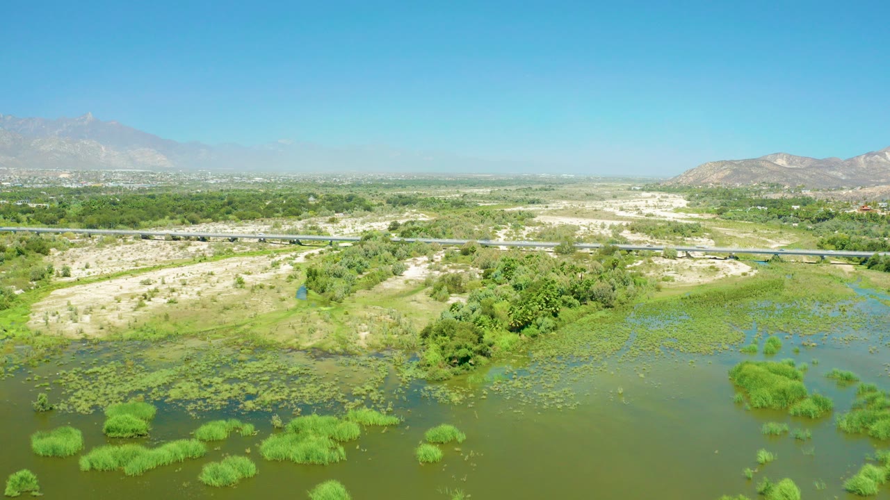 Aerial view of wetland water, reeds, bridge, dry land, mountains, and blue sky.