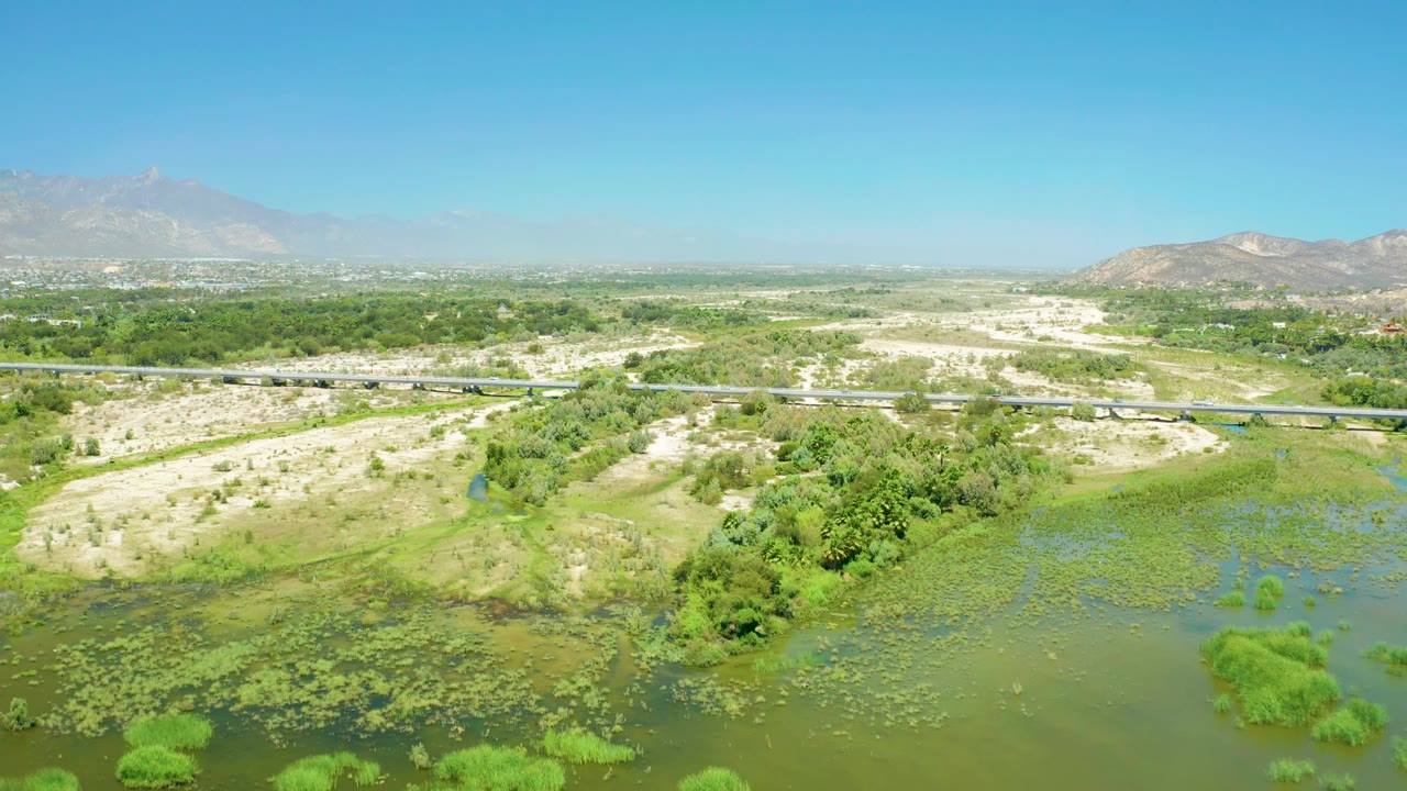 Mid-clip aerial frame over San Jose del Cabo Estuary where the green wetland water has receded lower in the composition, exposing more sandy terrain, trees, bridge, mountains, and no visible people.
