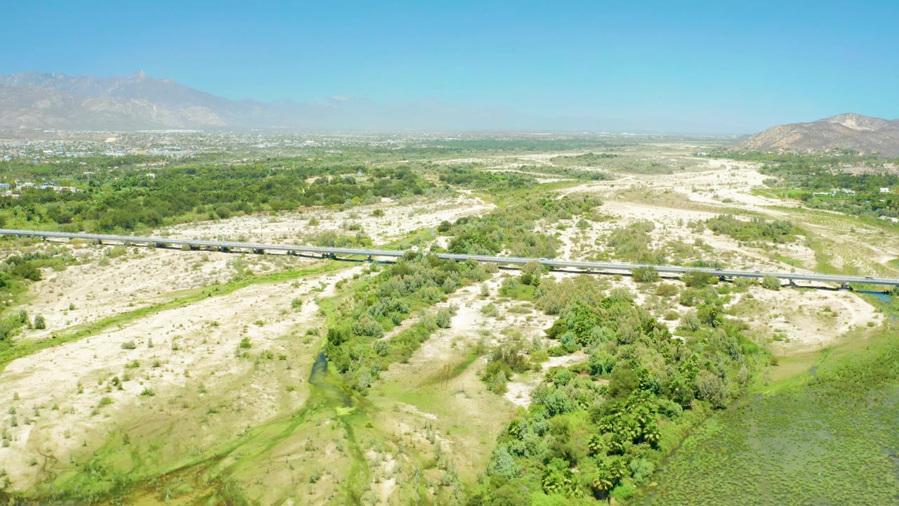 San Jose del Cabo Estuary aerial frame with the train continuing across the bridge, green trees and scrub below, pale open terrain to the right, mountains in the distance, and no visible people.