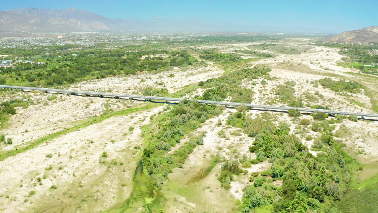 Wide aerial view of the San Jose Del Cabo Estuary area with a highway crossing pale sandy ground, green vegetation below, distant mountains, and no visible people.