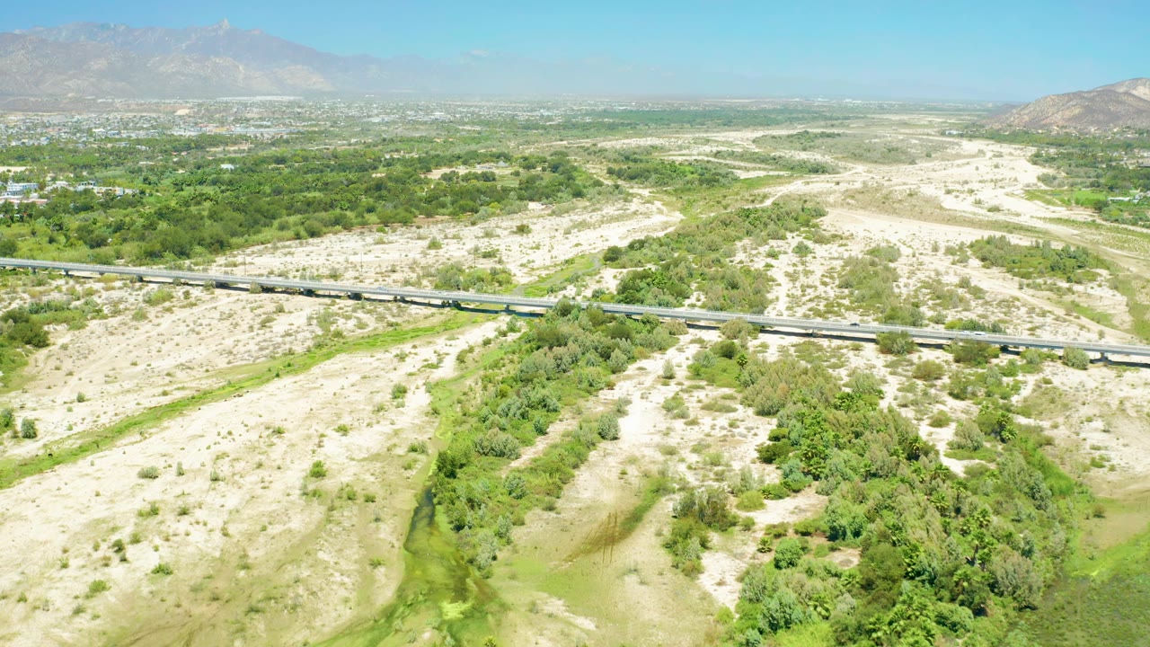 Aerial frame over San Jose Del Cabo Estuary showing the road angled across the midground, dry pale washes, a dense green channel, distant development, and no visible people.