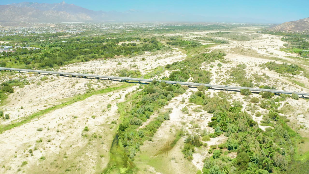 High drone view in San Jose Del Cabo Estuary with small vehicles on the highway, pale desert terrain on both sides, a green riparian strip below, mountains in haze, and no visible people.