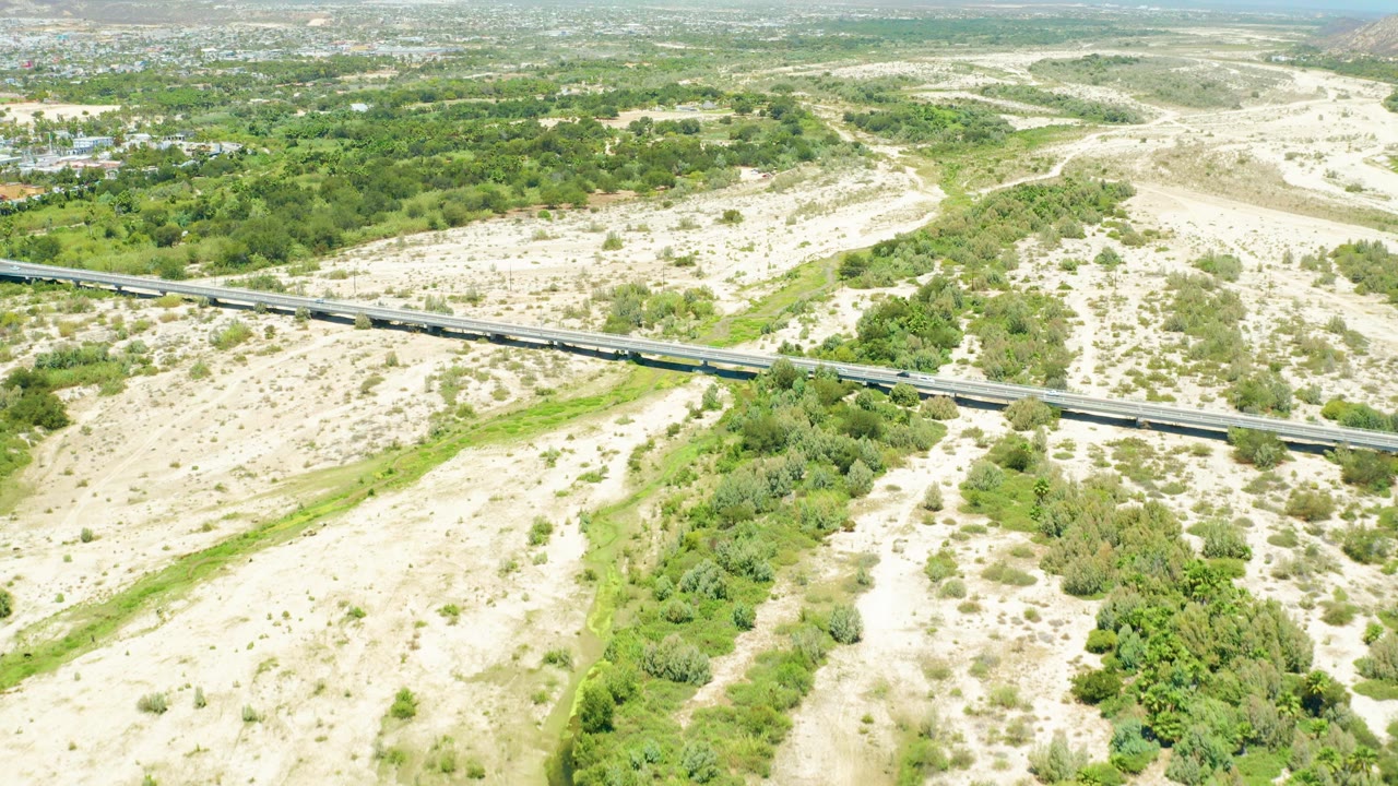 Aerial frame from San Jose Del Cabo Estuary showing a train crossing a bridge above pale sandy ground, green channel vegetation, and no visible people.