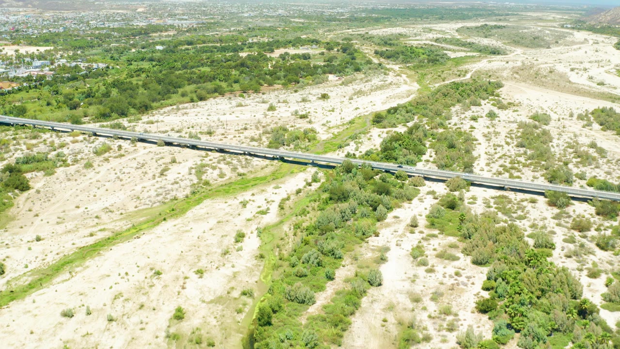 Aerial view in San Jose Del Cabo Estuary with the train slightly farther along the bridge, surrounded by sandy wash areas and scattered green vegetation, with no visible people.
