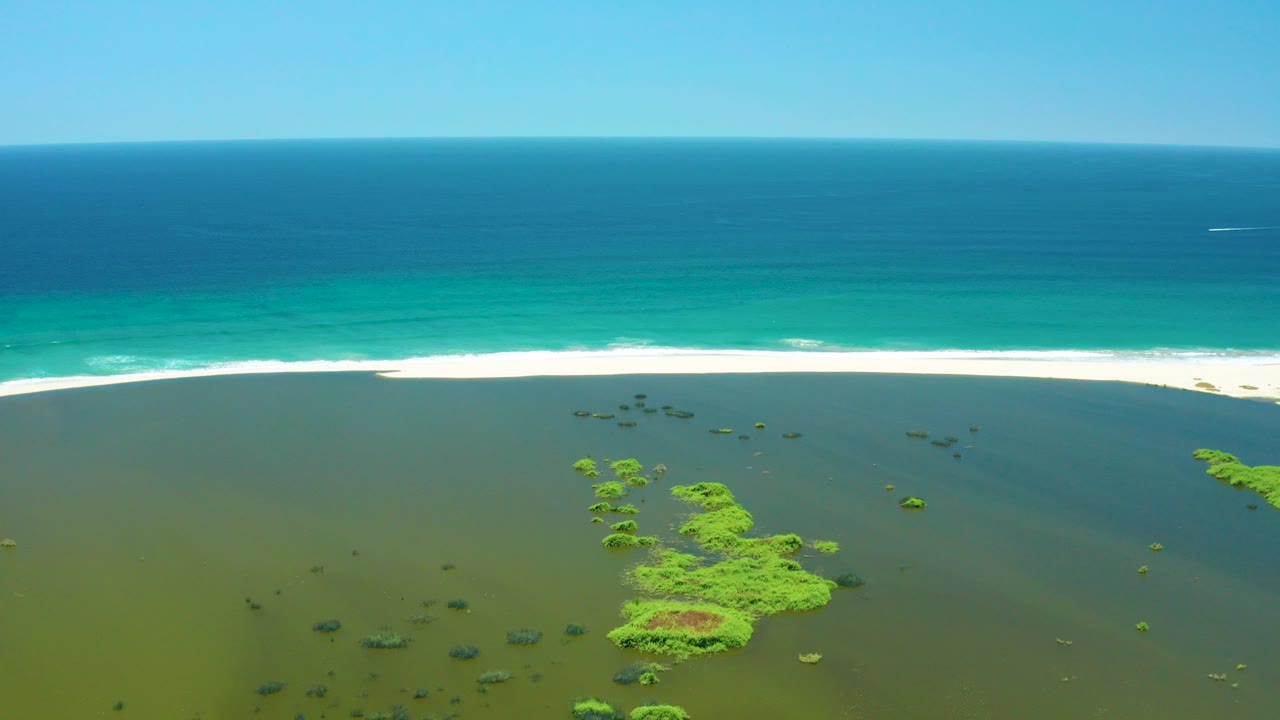 Opening aerial frame of San Jose Del Cabo Estuary with green wetland islets below, a pale beach and white surf line beyond, turquoise ocean, blue sky, and no visible people.