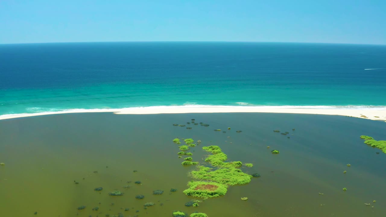 San Jose Del Cabo Estuary aerial frame with the main green islet cluster slightly lower in view, lagoon water in front, a continuous white wave band at the beach, and no visible people.