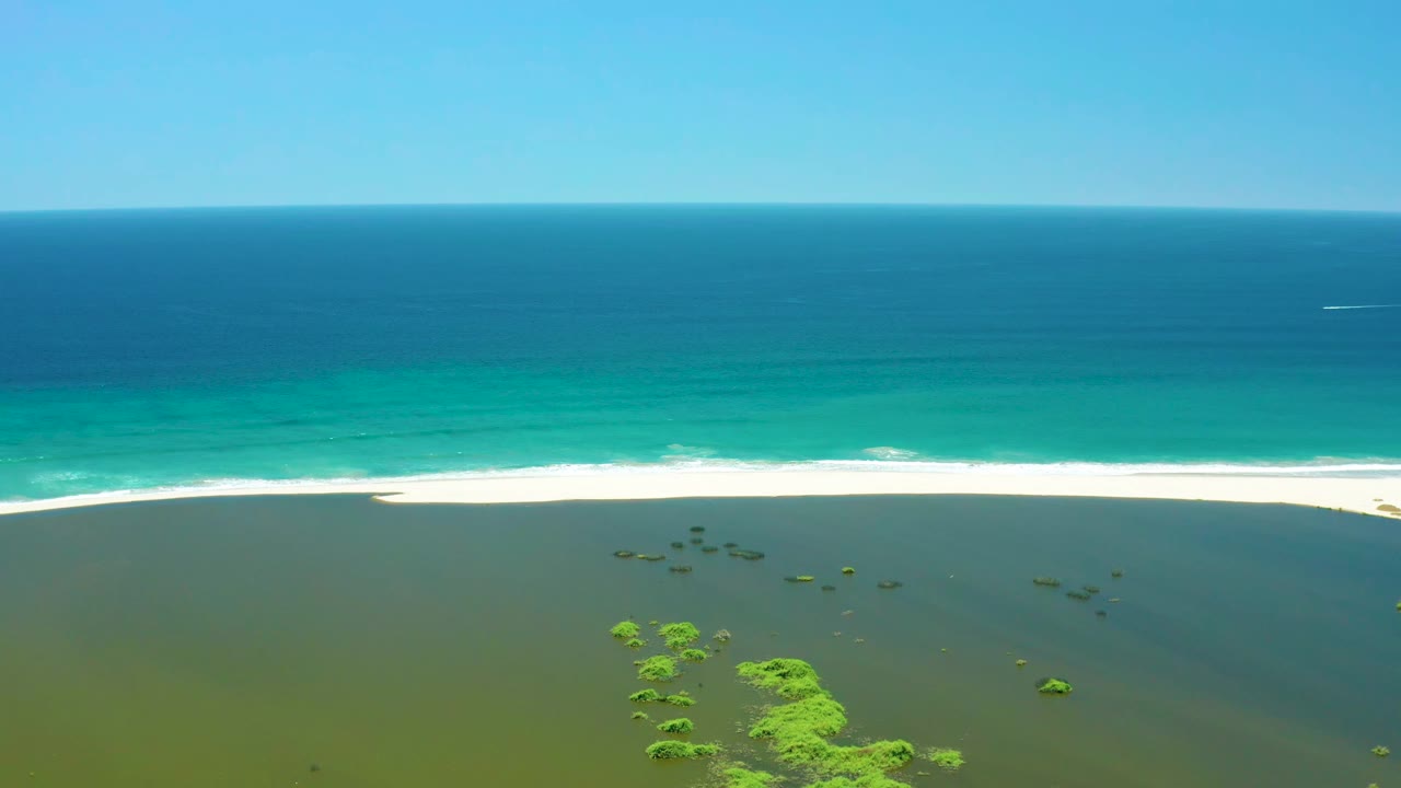 Aerial view over the San Jose Del Cabo Estuary with scattered green vegetation lower in frame, white surf tracing the beach, a small white wake offshore, and no visible people.