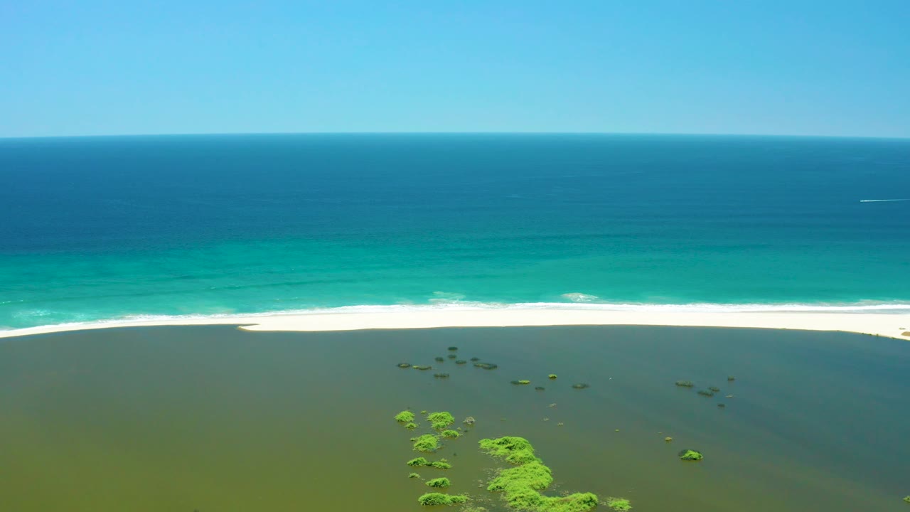 San Jose Del Cabo Estuary appears from above with green lagoon shallows in front, clustered vegetation near the lower center, a continuous white wave line on sand, open ocean, and no visible people.