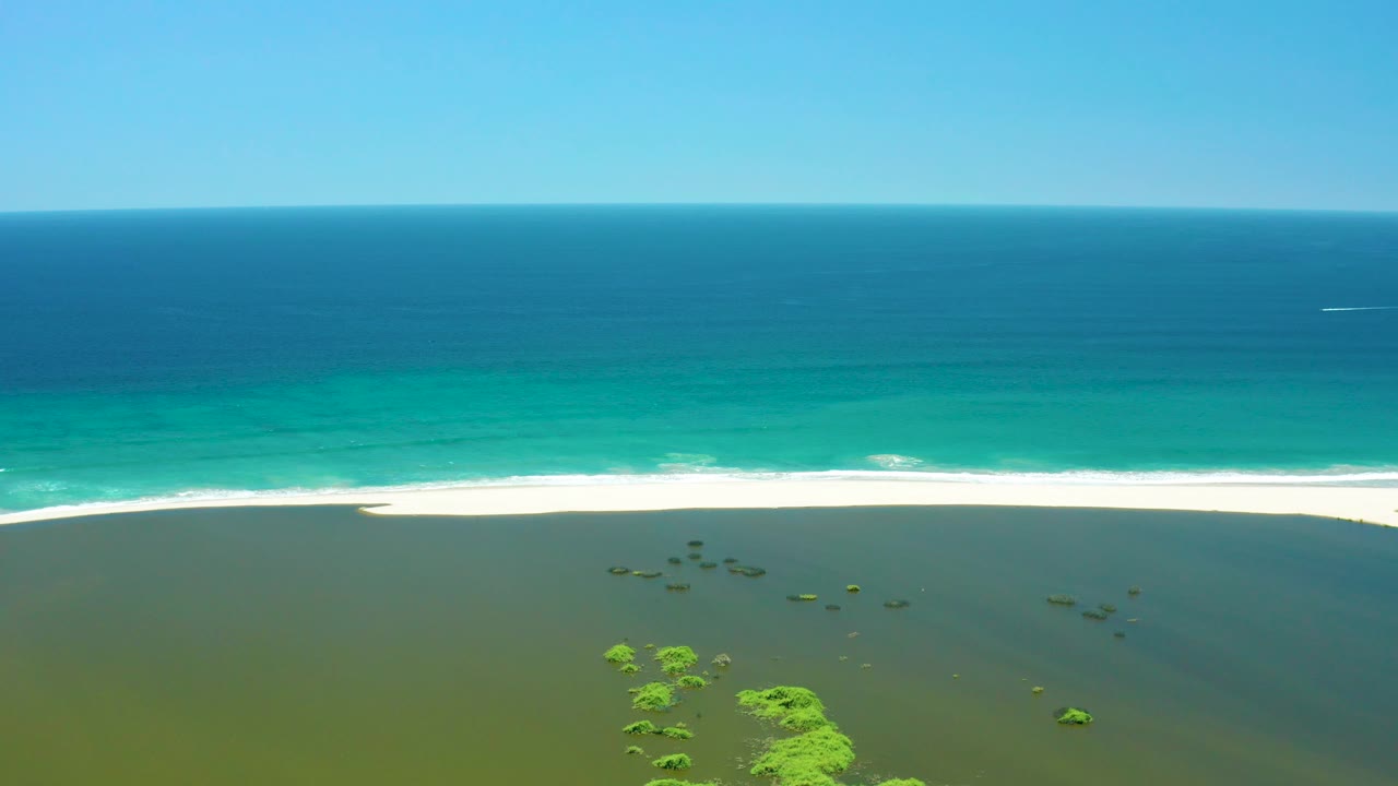 A wide aerial moment over San Jose Del Cabo Estuary shows the ocean horizon high in frame, a white surf band crossing the sand, darker green lagoon water below, and no visible people.