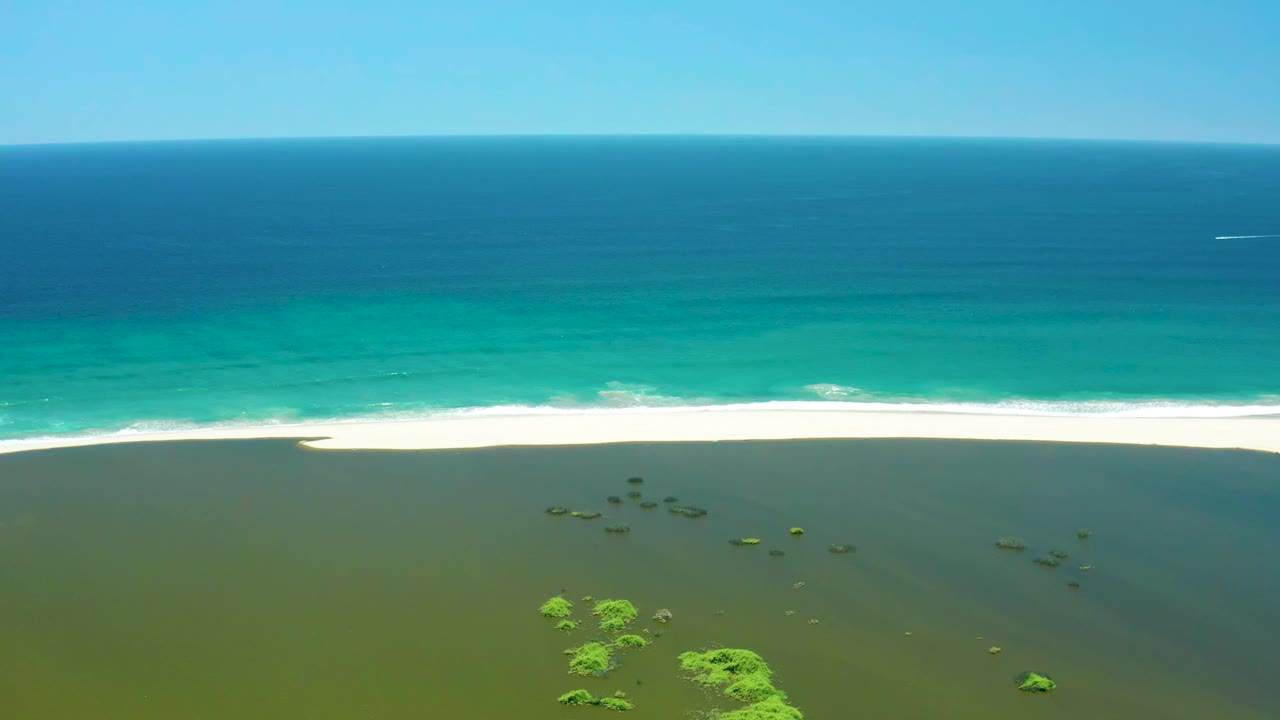 Aerial frame of the San Jose Del Cabo Estuary coast showing a clean white wave band along the sand, blue-green ocean beyond, green shallow water below, and no visible people.
