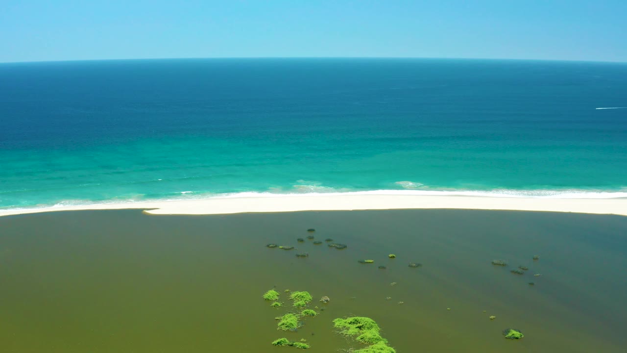 Wide aerial view at San Jose Del Cabo Estuary with the ocean filling the upper frame, a curved white surf line at midframe, vegetated green shallows below, and no visible people.