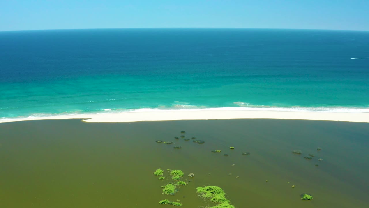 Aerial shoreline frame from San Jose Del Cabo Estuary with white waves breaking on pale sand, layered turquoise and blue water offshore, green shallows in front, and no visible people.