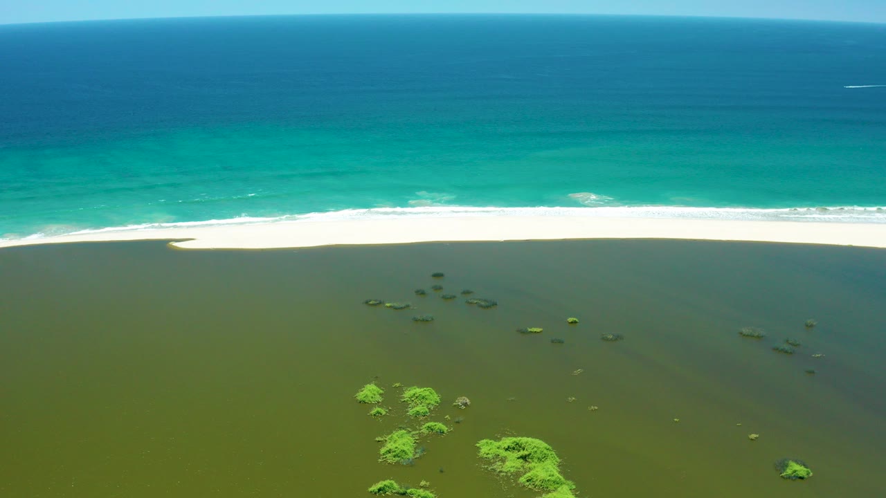 Opening aerial frame over San Jose Del Cabo Estuary, with deep blue ocean, turquoise surf, a pale sand strip, shallow green-brown wetland water, scattered vegetation, and no visible people.
