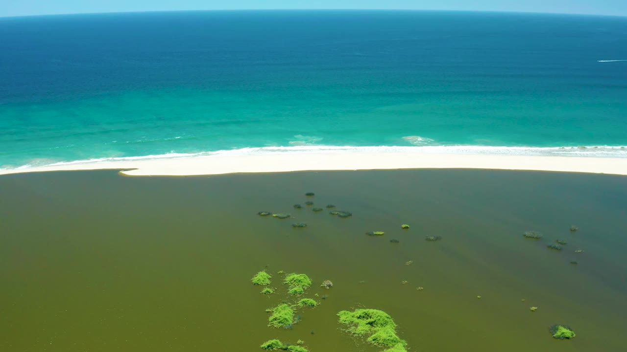 Aerial view at San Jose Del Cabo Estuary showing a long white surf line along the sandy beach, shallow wetland water in the foreground, small green vegetation clusters, and no visible people.