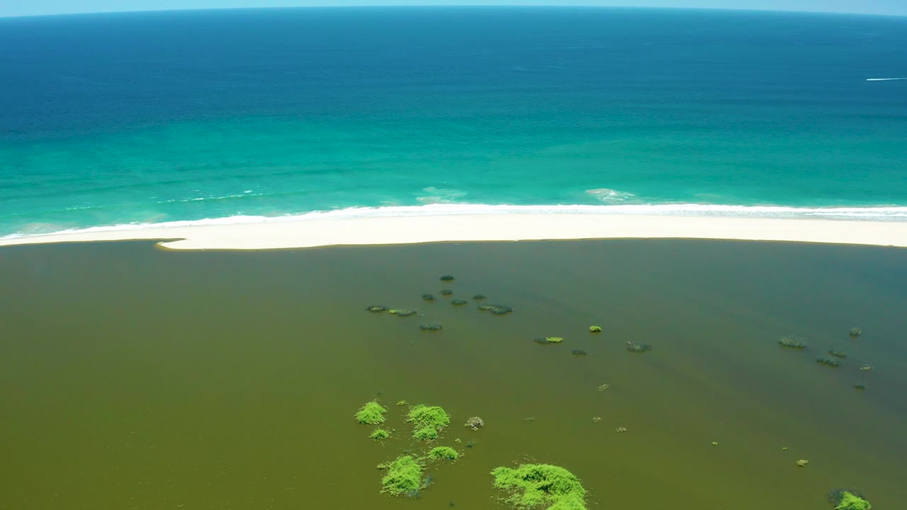 Aerial shot of ocean waves breaking beside a sandbar and shallow wetland water.