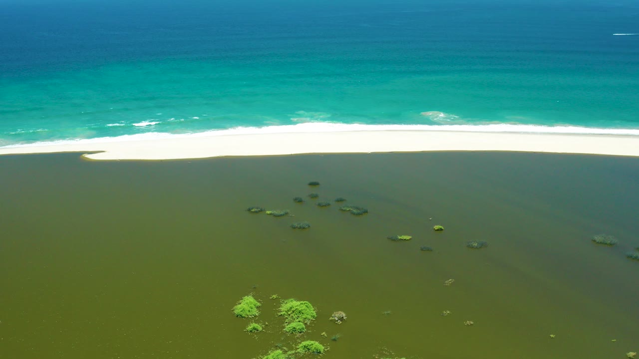Aerial view of turquoise ocean, white surf, green shallows, rocks, and mangrove patches.