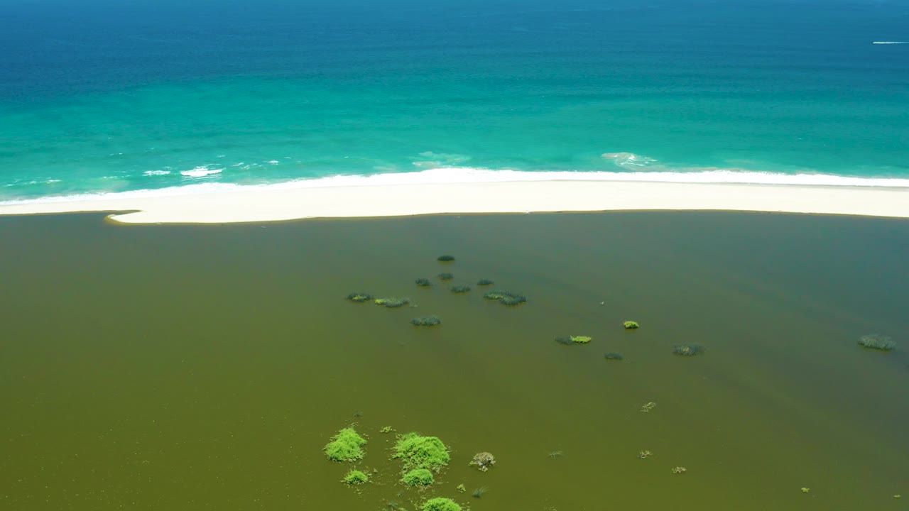 Aerial view of San Jose del Cabo Estuary with the ocean occupying the upper frame, a continuous white beach band across the middle, green shallow water below, rock clusters, mangrove patches, and no visible people.