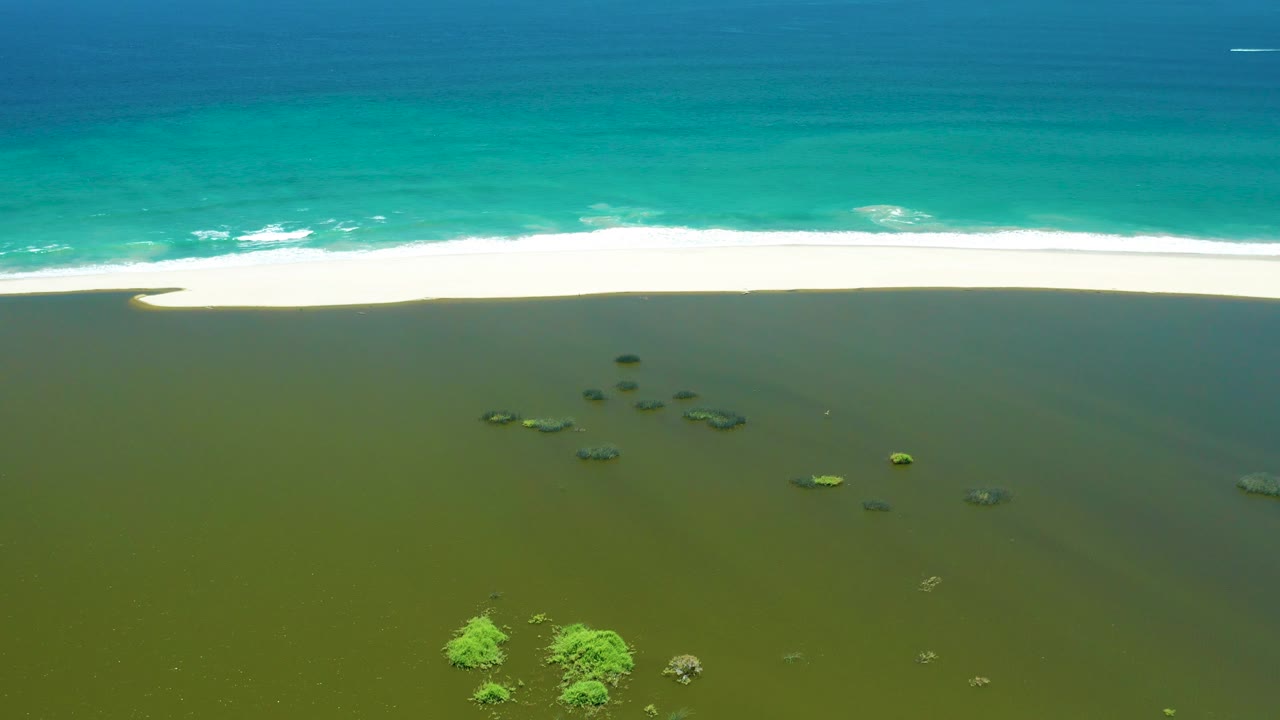 Mid-clip aerial frame over San Jose del Cabo Estuary where foamy turquoise surf presses toward the pale sand, green shallows fill the lower half, mangrove tops sit near the bottom edge, and no visible people appear.