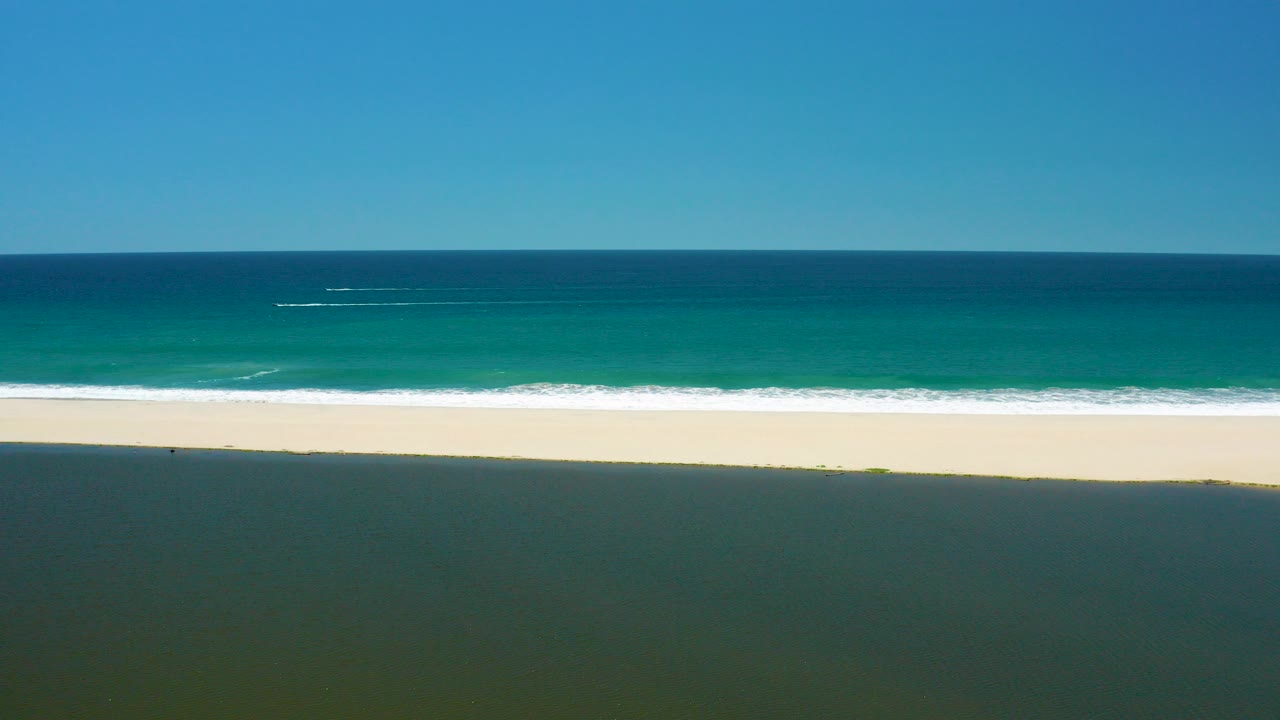 Opening frame of San Jose del Cabo Estuary shoreline with dark foreground water, a bright sand strip, turquoise ocean, clear sky, and no visible people.