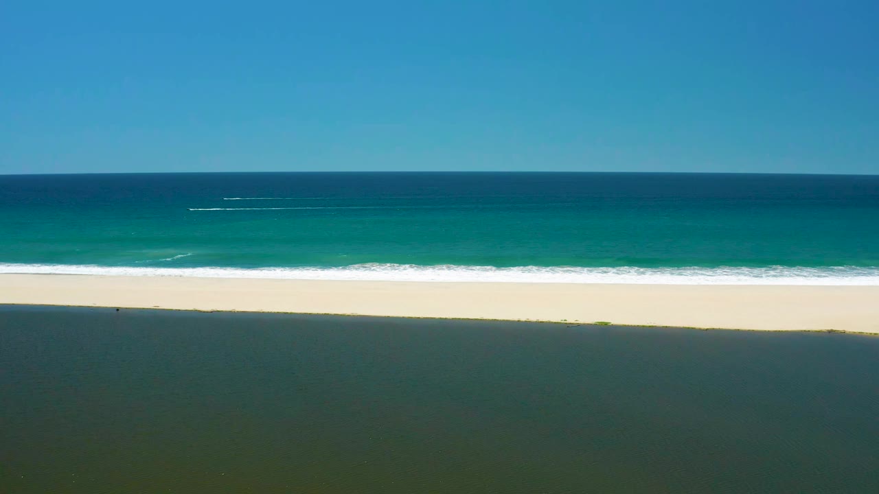 San Jose del Cabo Estuary coast at 0:01, showing a long white surf line against pale sand with blue-green ocean beyond and no visible people.