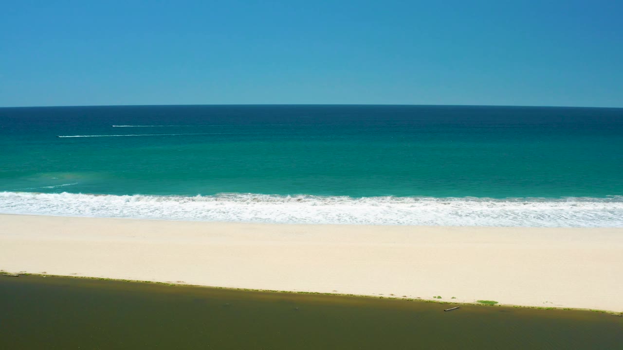 At 0:02, white foam stretches in a long horizontal band across the San Jose Del Cabo Estuary shoreline, backed by turquoise and deep blue water with no visible people.