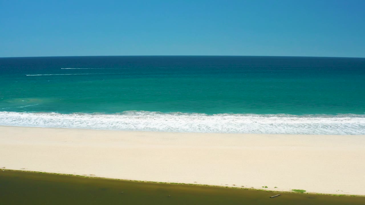 At 0:03, a darker strip of coastal vegetation or shadow borders the lower edge while pale sand, foamy surf, turquoise water, and open sky fill the San Jose Del Cabo Estuary view with no visible people.