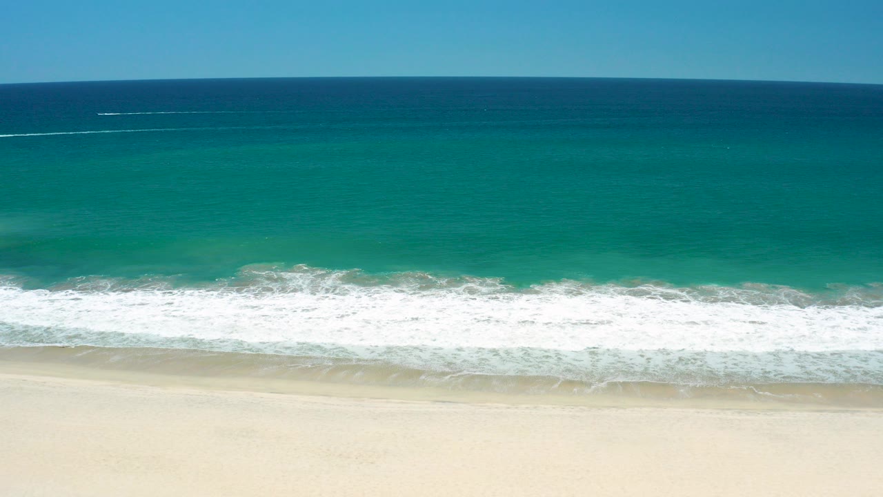 Wide aerial frame over San Jose Del Cabo Estuary shoreline, with deep blue ocean near the horizon, turquoise water, white surf, pale sand, and no visible people.