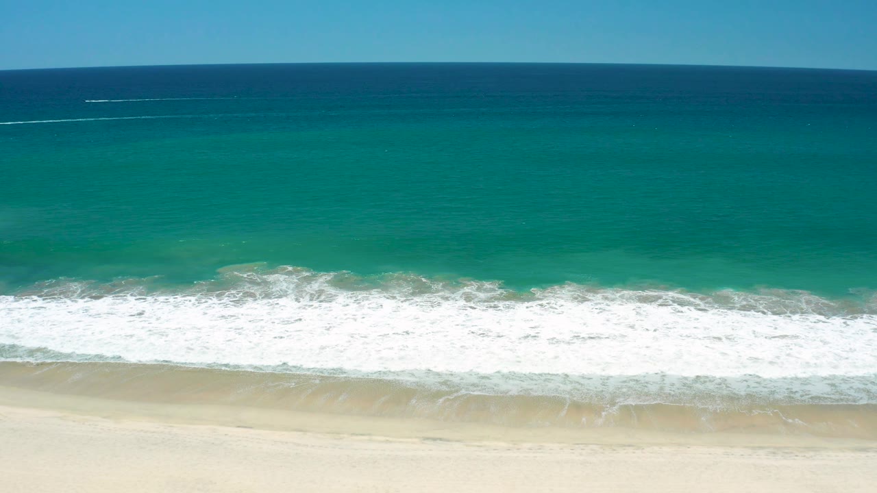 San Jose Del Cabo Estuary shoreline from above, with deeper blue water at the top, turquoise shallows below, a foamy wave edge, sand at the bottom, and no visible people.