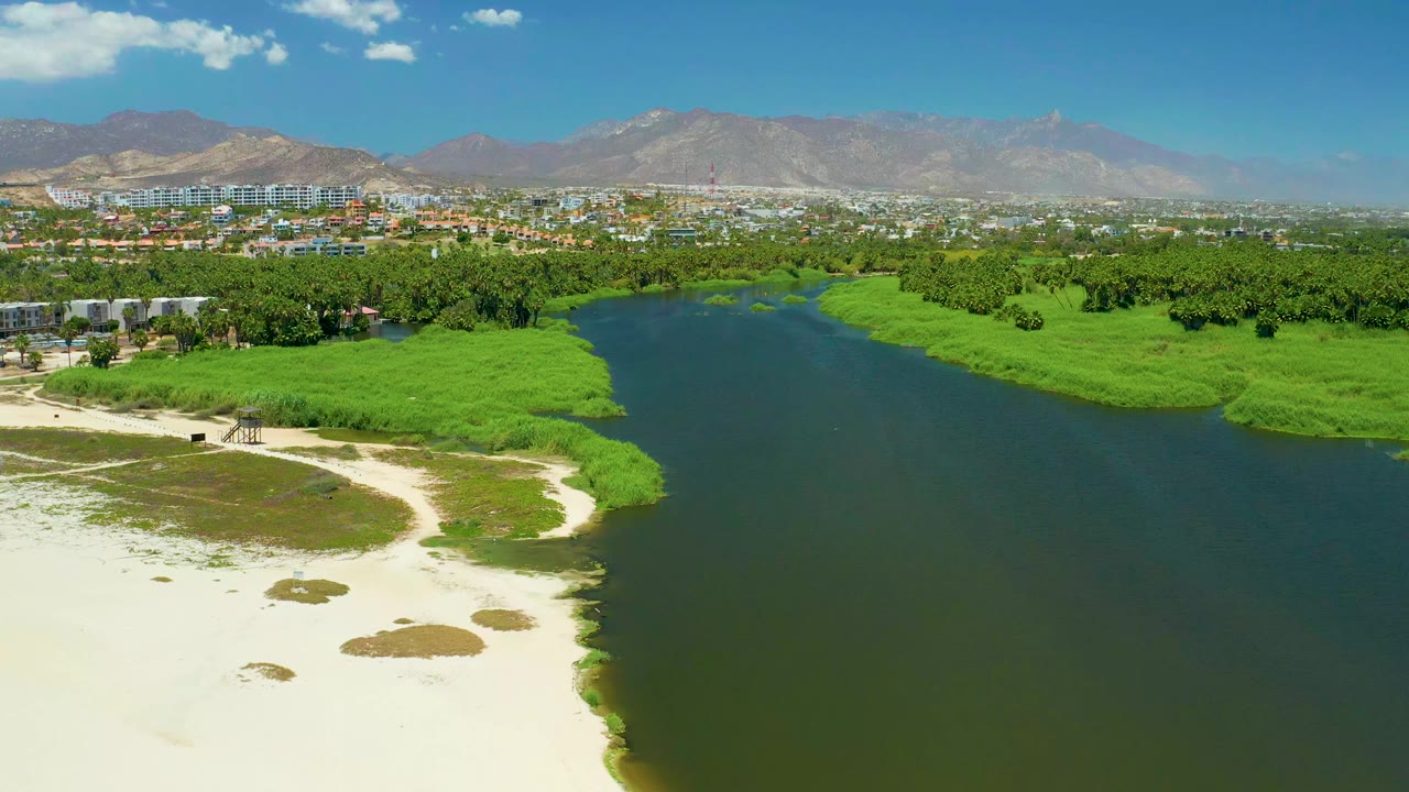 Wide aerial frame of the San Jose del Cabo Estuary showing the river channel centered between marsh plants, with sandy shoreline at lower left, distant buildings, mountains, and no visible people.